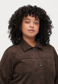 Giacca in ecopelle scamosciata marrone con collo a camicia, due bottoni neri e una texture liscia; presenta un design semplice senza motivi.