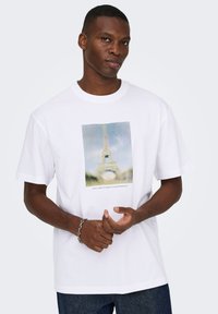 Λευκό βαμβακερό μπλουζάκι με κοντά μανίκια και άνετη εφαρμογή, στο κέντρο του οποίου υπάρχει μια γραφική εικόνα του Πύργου του Άιφελ με θολό φόντο.