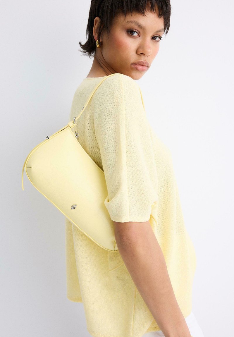 Femme portant un haut texturé jaune pâle avec un sac à main jaune assorti à l'épaule, sur fond blanc.