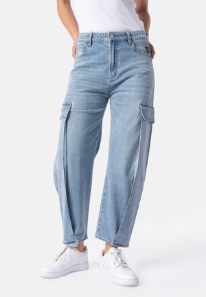 Person iført lyseblå denim cargobukser og hvide sneakers, stående med hænderne i lommerne mod en ensfarvet baggrund.
