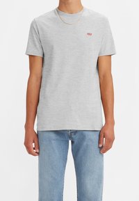 T-shirt grigio a maniche corte in cotone con colletto rotondo, con un piccolo logo rosso di Levi's sul lato sinistro del petto. Indossato con jeans azzurri chiari.
