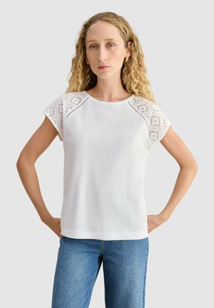 Mujer con cabello rubio largo y rizado, que lleva una camiseta blanca con mangas de encaje y vaqueros azules, de pie con las manos en las caderas.