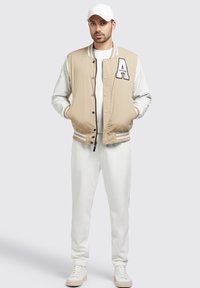 Jachetă de tip varsity bej cu mâneci albe, având un patch cu litera "A", nasturi negri și manșete cu dungi, asortată cu pantaloni jogger albi și adidași.