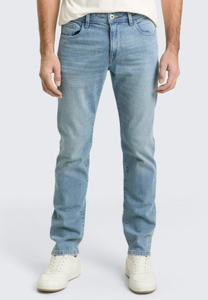 Man draagt lichtblauwe rechte spijkerbroek en witte sneakers, staand tegen een effen lichte achtergrond.