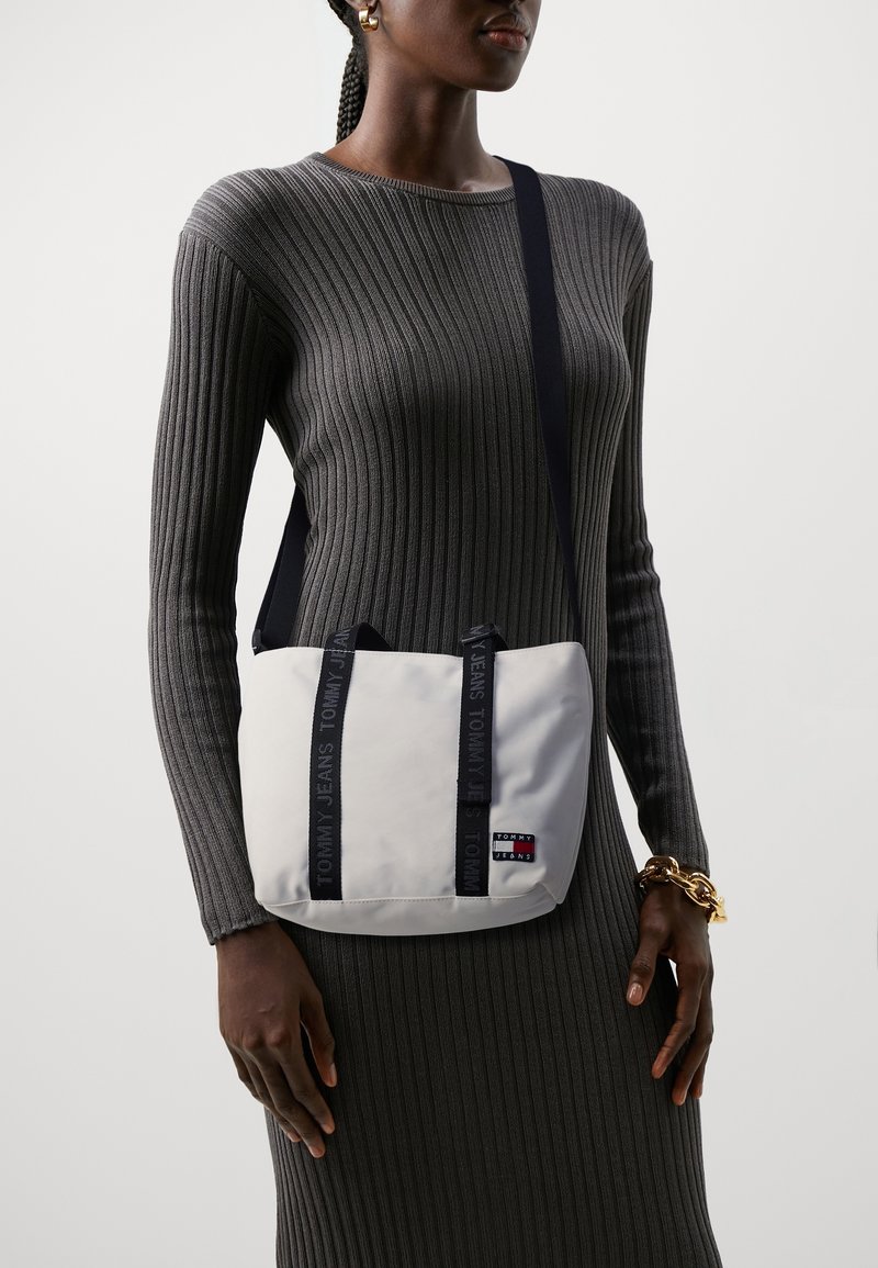 Femme portant une robe grise côtelée avec un sac bandoulière blanc Tommy Jeans et un bracelet chaîne doré au poignet.