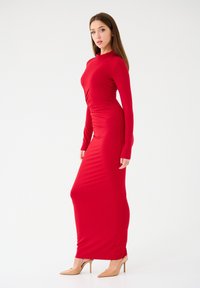 Abito maxi rosso a maniche lunghe in tessuto elasticizzato, con una silouette aderente e dettagli arricciati. Accompagnato da tacchi alti in tonalità neutra.