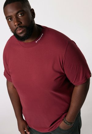 Bordeauxfarbenes T-Shirt mit kurzen Ärmeln, Rundhalsausschnitt und Logo auf dem Kragen. Glatte Textur, lässige Passform, kombiniert mit einer dunklen Jeans und Armbändern.
