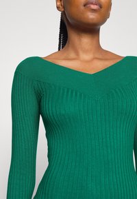 Zelený potrhaný dlouhý rukáv s výstřihem do V. Obsahuje vertikální pruhy a přiléhavý střih, který zvýrazňuje kontury těla.