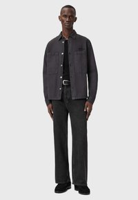 Uomo alto che indossa una camicia grigia scura con bottoni sopra una maglietta nera, con cintura nera, jeans neri e scarpe nere, in piedi di fronte a uno sfondo grigio.