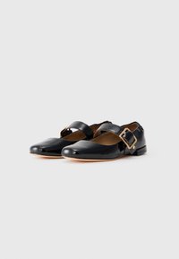 Bailarinas de charol negro tipo Mary Jane con correas anchas y grandes hebillas en tono dorado, suelas en beige neutro y tacones bajos.