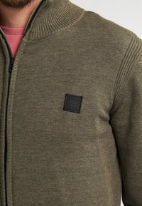 Olivgrüner Strickreißverschluss-Pullover mit geripptem Kragen und Akzentpanel. Verfügt über ein kleines schwarzes quadratisches Logo-Patch auf der linken Seite.