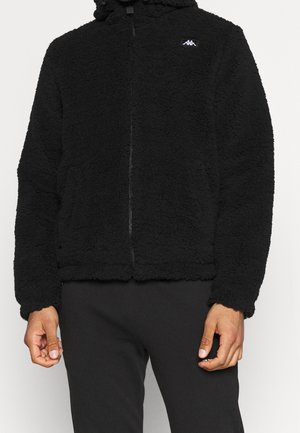 Schwarze Fleecejacke mit Reißverschluss und hohem Kragen, strukturierter Stoff und kleines weißes Logo auf der Brust, getragen mit schwarzer Hose.
