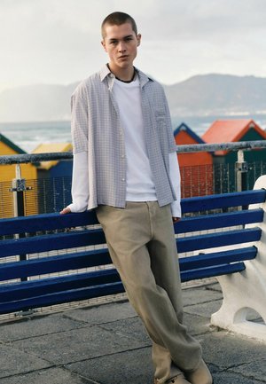 Junger Mann lehnt an einer blauen Bank an der Strandpromenade, trägt ein weißes Langarmshirt unter einem karierten Kurzarmhemd und beige Hose.