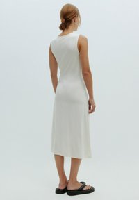 Femme aux cheveux courts bruns portant une robe midi blanche sans manches et des sandales à plateforme noires, debout devant un fond clair uni.