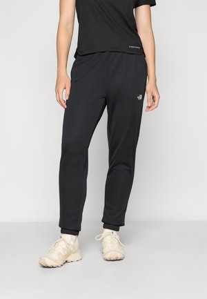 Personne portant un pantalon jogging noir et un haut à manches courtes noir, debout avec des chaussures de sport crème à lacets sur un sol gris.