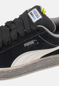 Tenisi negri din suede cu accente crem, talpă din cauciuc texturată și cusături verzi. Prezintă un logo Puma proeminent și șireturi negre.