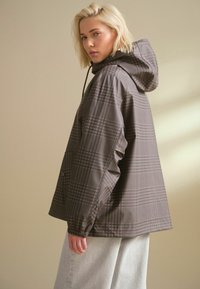 Chaqueta de lluvia a cuadros grises hecha de tela resistente al agua, con capucha, puños elásticos y bolsillos frontales para mayor funcionalidad.