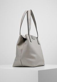Sac fourre-tout en cuir beige avec une finition texturée, deux poignées supérieures et une fermeture éclair verticale. Quincaillerie argentée et accent logo minimaliste.