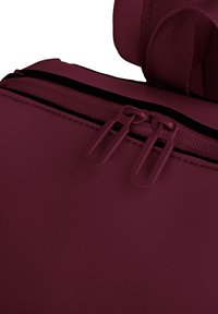 Sac à dos bordeaux foncé avec une texture lisse et mate ; il dispose de doubles tirettes de fermeture éclair et d'une bandoulière rembourrée.