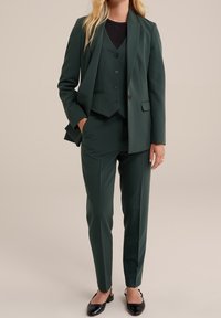 Grön tre-delad kostym bestående av en figursydd blazer, väst med knappar och avsmalnande byxor. Materialet verkar slätt med en matt yta.