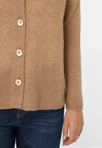 Cardigan in maglia marrone con vestibilità rilassata, dotato di grandi bottoni beige, polsini a coste e un accento laterale vicino all'orlo.