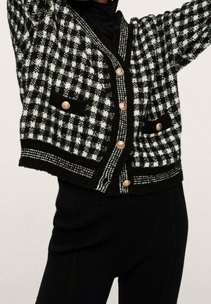 Cardigan noir et blanc à motif pied-de-poule avec des boutons dorés et deux poches avant, porté sur une tenue noire côtelée.