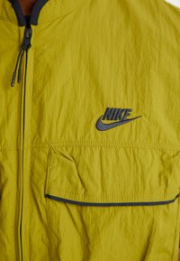 Nike striukė ryškiai geltonos spalvos, turinti užtrauktuką, tekstūruotą medžiagą, juodą siuvinėjimą ir mažą juodą logotipą kairiajame viršutiniame kampe.