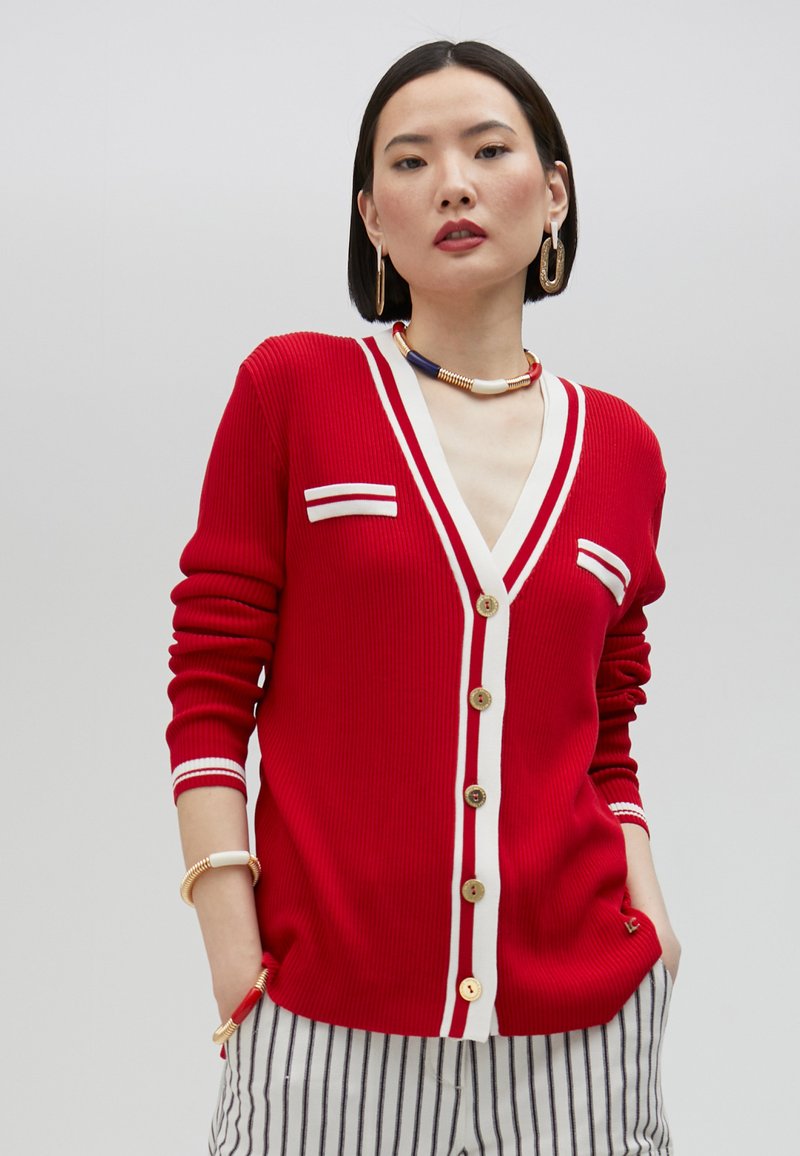 Red ribbed cardigan with white trim and two small front pockets, featuring gold buttons and a V-neckline. Striped pants visible below.