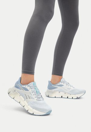 Person trägt graue Leggings und weiße-blau Reebok Laufschuhe und steht vor einem weißen Hintergrund.