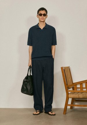 Homme portant un pull en maille bleu marine à manches courtes, un pantalon large foncé, des sandales et des lunettes de soleil, tenant un sac en cuir noir, debout à côté d'une chaise en bois.