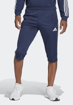 Mann trägt marineblaue Adidas Dreiviertel-Trainingshose mit weißen Streifen und weißen Sportschuhen, steht vor hellem Hintergrund.
