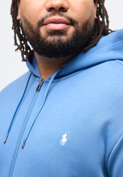 Άνδρας με γένια που φοράει μπλε φούτερ με φερμουάρ και λευκό λογότυπο παίκτη πόλο στο στήθος, εμφανίζοντας τον άνω κορμό και το κάτω μέρος του προσώπου.