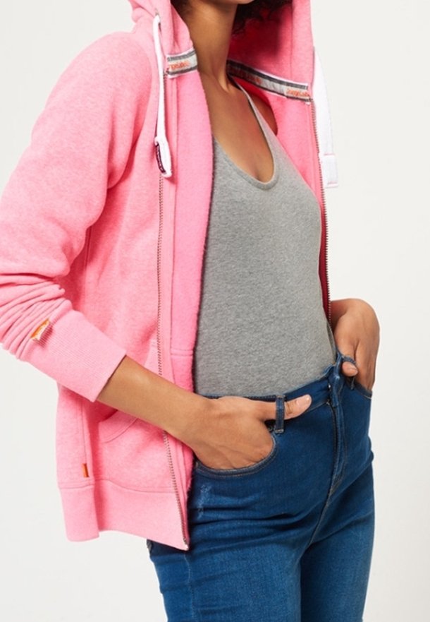 Sudadera rosa con cremallera y capucha ajustable, forro interior en contraste y bolsillos, llevada sobre una camiseta sin mangas gris y jeans azules.