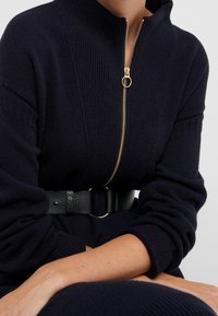 Marinblå stickad klänning med hög krage och detaljer i guldfärgad dragkedja. Har ett svart strukturerat bälte, framhäver taperade ärmar och en figursydd silhuett.