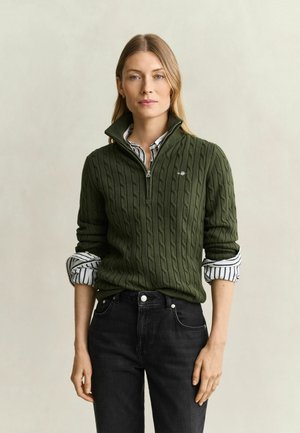 Frau mit glattem blondem Haar trägt einen olivgrünen Grobstrickpullover über einem gestreiften Hemd mit hochgekrempelten Ärmeln und schwarzen Jeans vor einem schlichten Hintergrund.