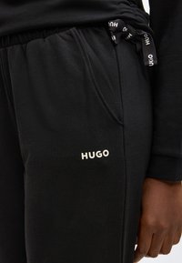 Črne trenirka hlače iz mehkega blaga, z bočnim žepom, elastičnim pasom in belim logotipom, vezenim na levi stegni.