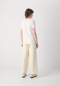 Hvit T-skjorte med korte ermer kombinert med løstsittende kremfargede bukser. Føttene har på seg beige sko. Minimalistisk, myk tekstur.