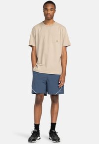 Beige Baumwoll-T-Shirt mit einer Brusttasche, kombiniert mit navyblauen Sportshorts mit weißen Nähten und einem Logo. Schwarze Sneakers runden das Outfit ab.
