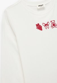 Biała koszula z długim rękawem z czerwoną haftowaną grafiką przedstawiającą prezent, wstążkę i pluszowego misia. Wykonana z miękkiego materiału z okrągłym dekoltem.