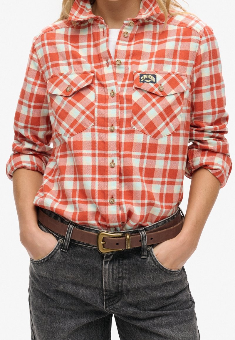 Chemise en flanelle à carreaux rouge et bleu clair avec deux poches poitrine, devant à boutons et manches retroussées. Portée avec un jean foncé et une ceinture marron.