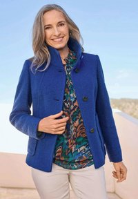 Blaue Wolljacke mit hohem Kragen, Knopfverschluss und zwei vorderen Taschen, getragen über einer gemusterten Bluse in Grün-, Orange- und Blautönen.