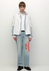 Camicia oversize a righe bianche sovrapposta a un polo azzurro chiaro, abbinata a jeans in denim larghi con testo grafico rosso e accenti a forma di stella. Scarpe nere.