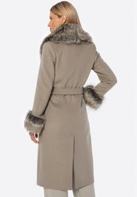 Beige vlněný kabát s kožešinovým límcem a manžetami, opasek v pase, dlouhé rukávy, rovný střih a délka do poloviny stehen. Měkká textura a čisté linie.