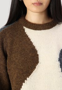 Brauner und cremefarbener Wollpullover mit geometrischen Mustern, ausgestattet mit strukturiertem Strick, einem runden Ausschnitt und dunkelgrauen Akzenten an den Schultern.