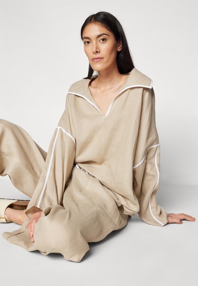 Ensemble en lin beige comprenant un haut ample à col blanc et à manches longues, accompagné d'un pantalon loose assorti, agrémenté de détails en piping blanc.