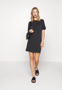 Vestido preto de mangas curtas com corte reto, feito de tecido suave. Combinado com sandálias de salto baixo pretas e uma pequena bolsa de ombro preta.