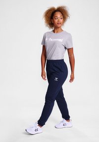 Graues T-Shirt mit "hummel"-Logo, marineblaue Jogginghose, weiße Sneakers, Strukturstoff, figurbetontes Design, elastischer Bund, Seitentaschen, gestickte details.