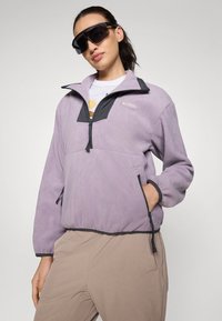 Svetlofialový fleece pulóver so zipsom na golieri, čiernymi akcentmi a dvoma bočnými vreckami. Na ľavej hrudi sa nachádza logo.