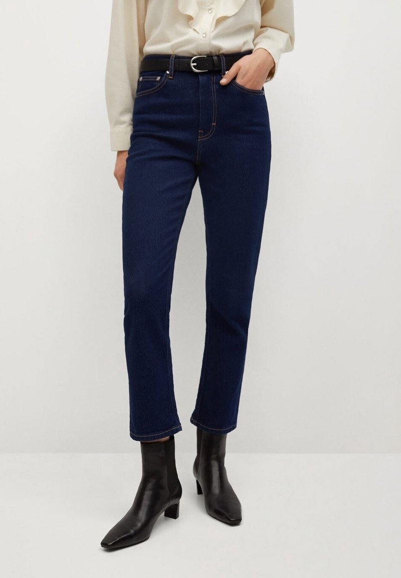 Femme portant un jean droit bleu foncé avec ceinture noire, blouse crème et bottines à talons noires, debout devant un fond blanc.