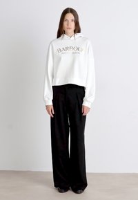 White cropped hoodie with "BARBOUR INTERNATIONAL" printed in gold, paired with black wide-leg trousers; smooth fabric, relaxed fit.